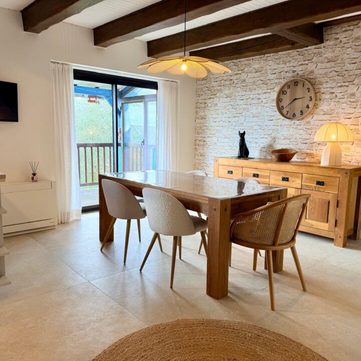 Salle à manger lumineuse avec table en bois, poutres apparentes et mur en pierre.