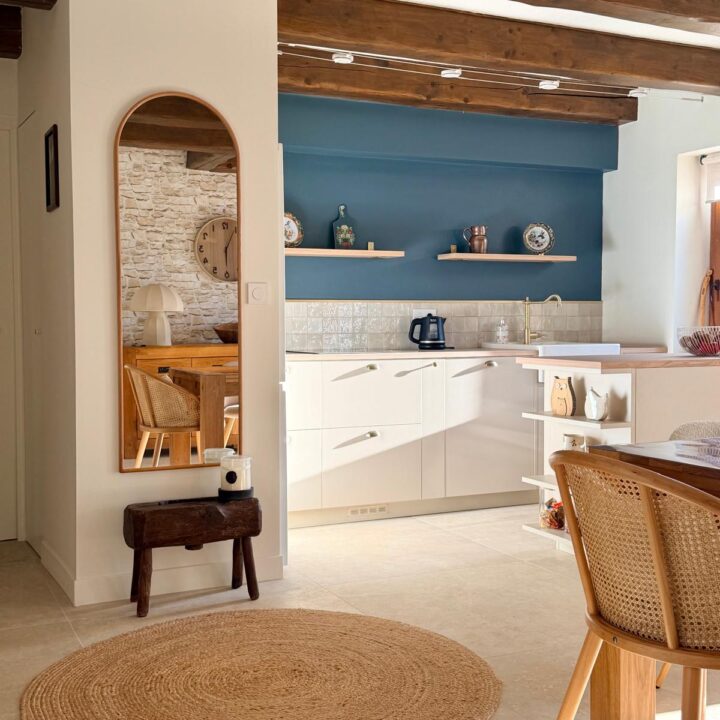 Vue sur une cuisine ouverte blanche avec mur bleu pétrole, poutres en bois et miroir arrondi.