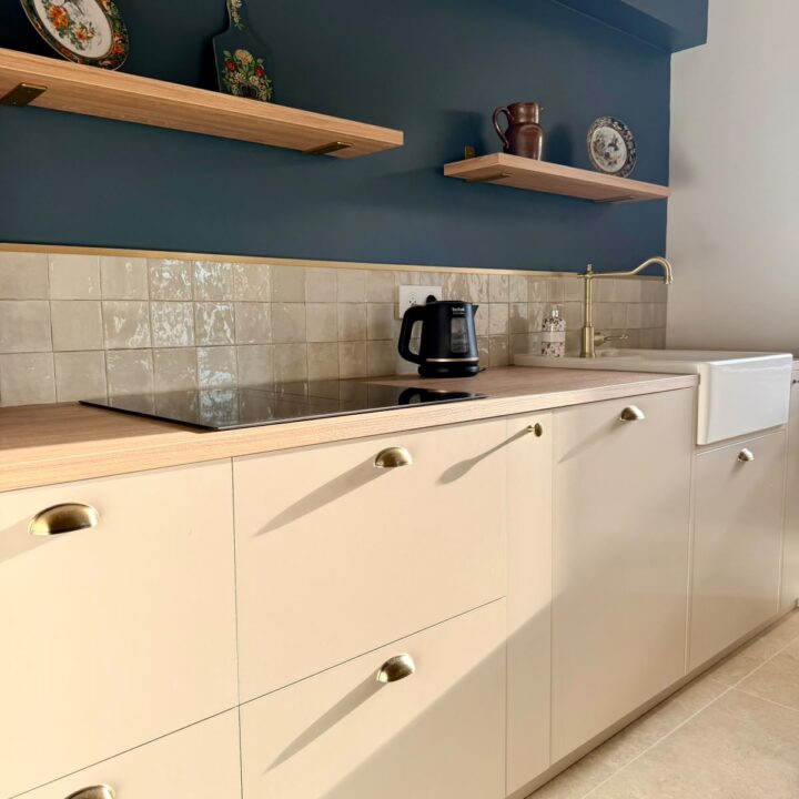 Détail d’une cuisine blanche avec mur bleu pétrole, étagères en bois et crédence en carrelage beige.